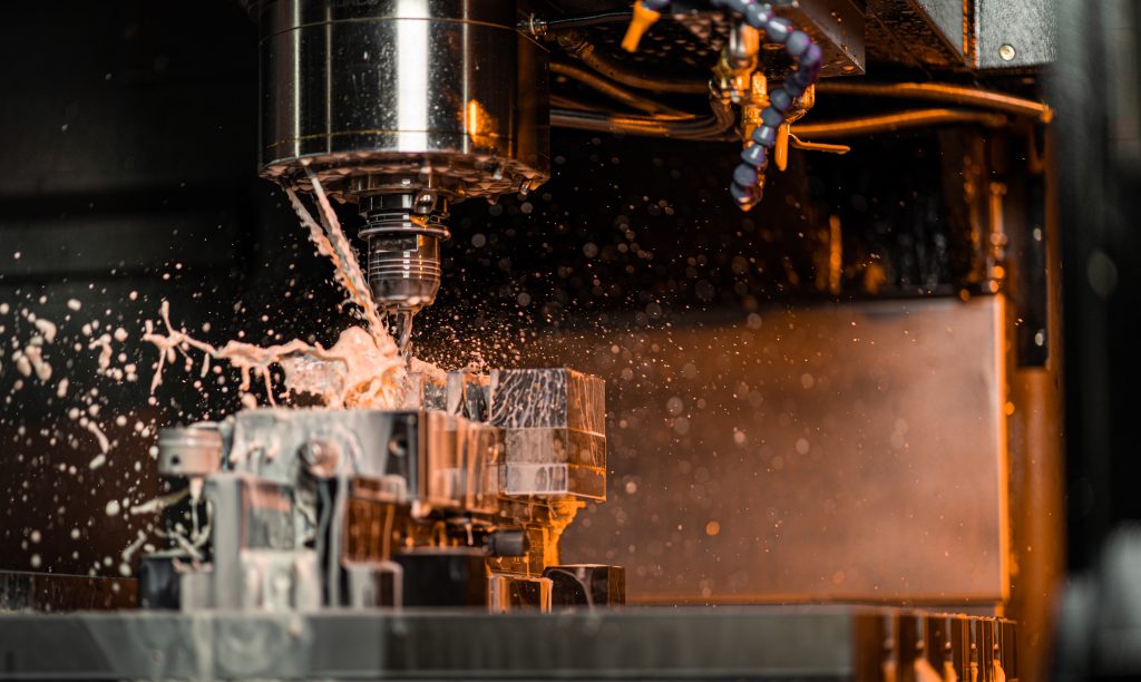 Close-up inside a CNC milling machine as coolant sprays around the cutting tool while machining a metal workpiece, with droplets and chips in motion.