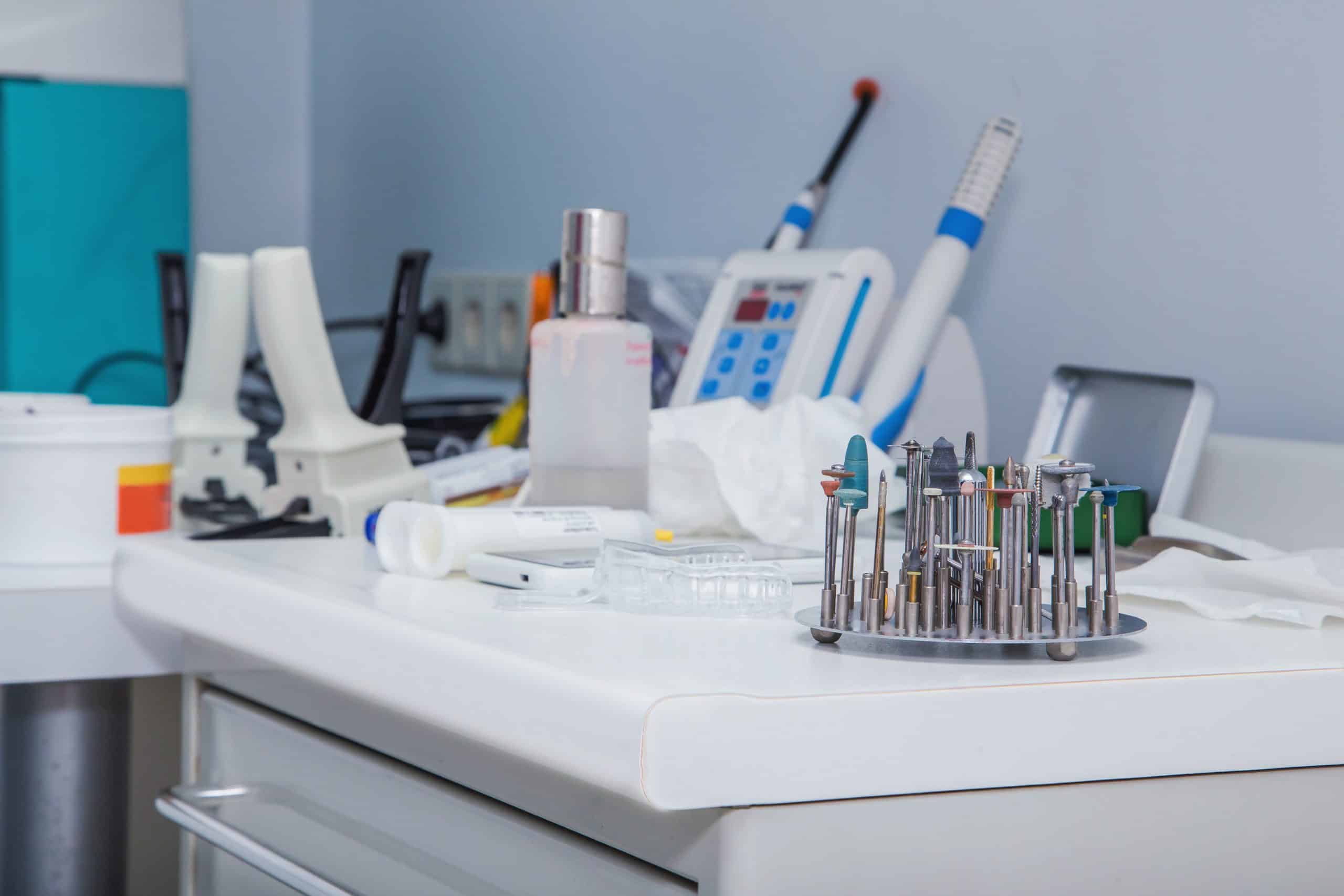 Close-up of a clinical workstation with rotary tools, handheld instruments, bottles, and medical supplies arranged on a white counter.